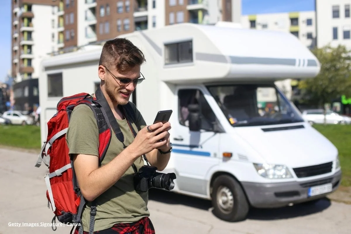 Tecnología y turismo sostenible en Colombia: innovación para un futuro responsable 1 Joven viajero con mochila y cámara usando un teléfono móvil frente a una autocaravana en ciudad de Colombia para turismo sostenible 2025.
